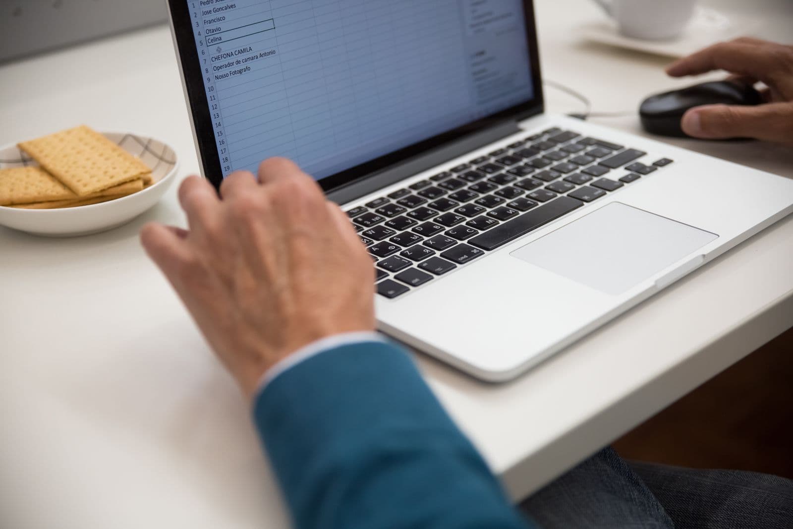 A laptop screen showing a spreadsheet with a column of names, one hand on the keyboard and one on a mouse