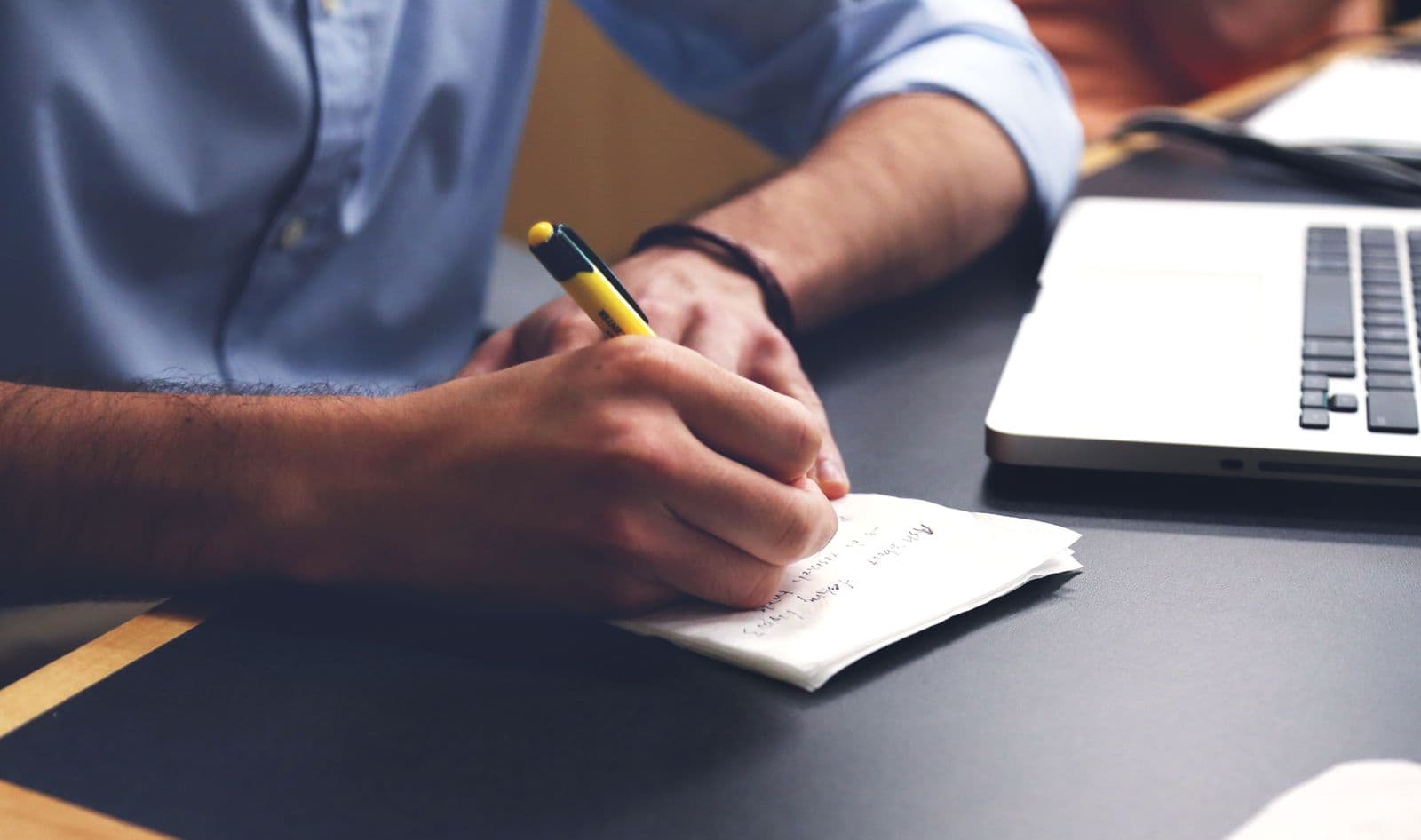A man writing notes on a folded sheet of paper with a yellow pen, an open laptop on the desk beside him