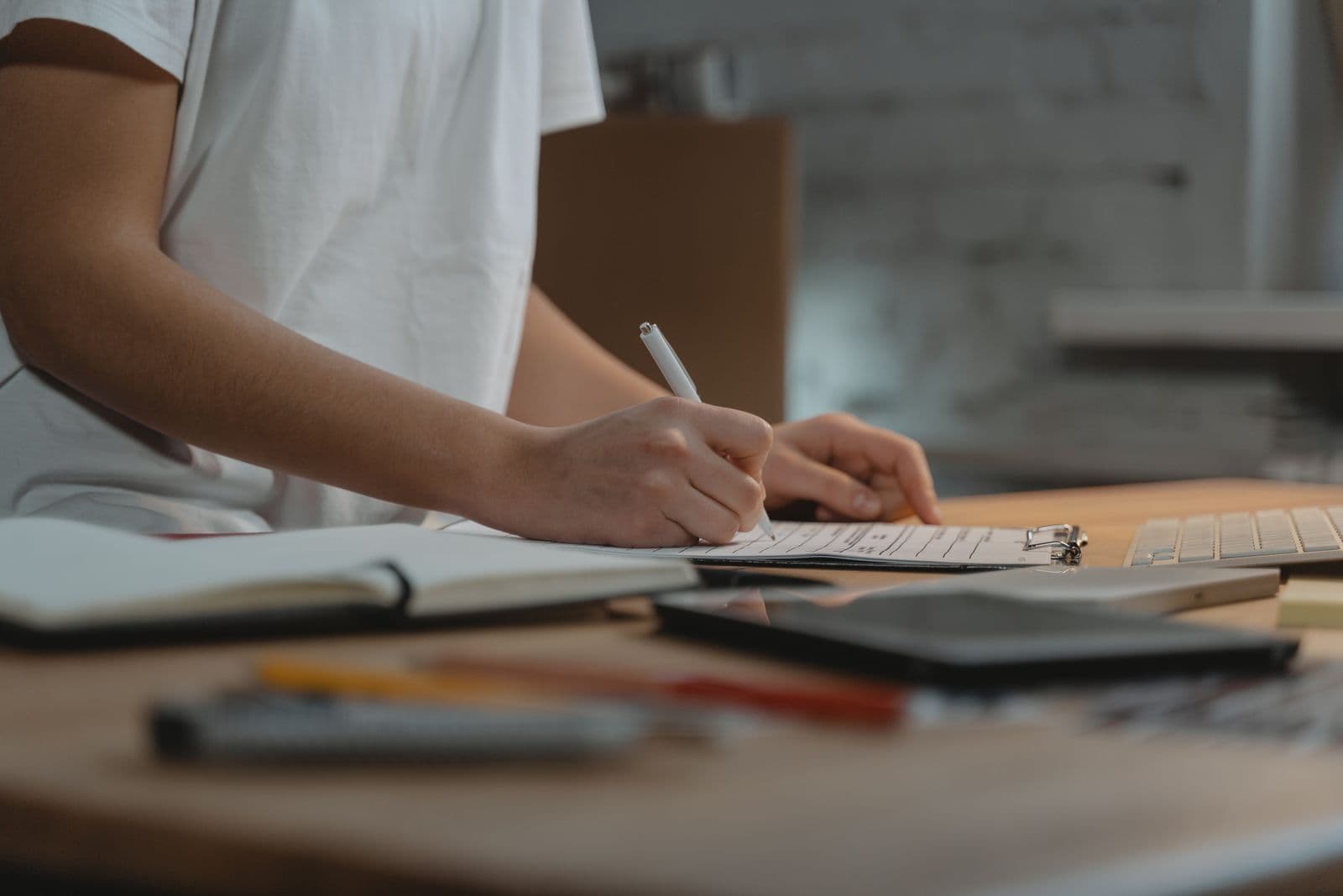 A person filling in a printed checklist on a clipboard, an open notebook, pens and a keyboard on the desk