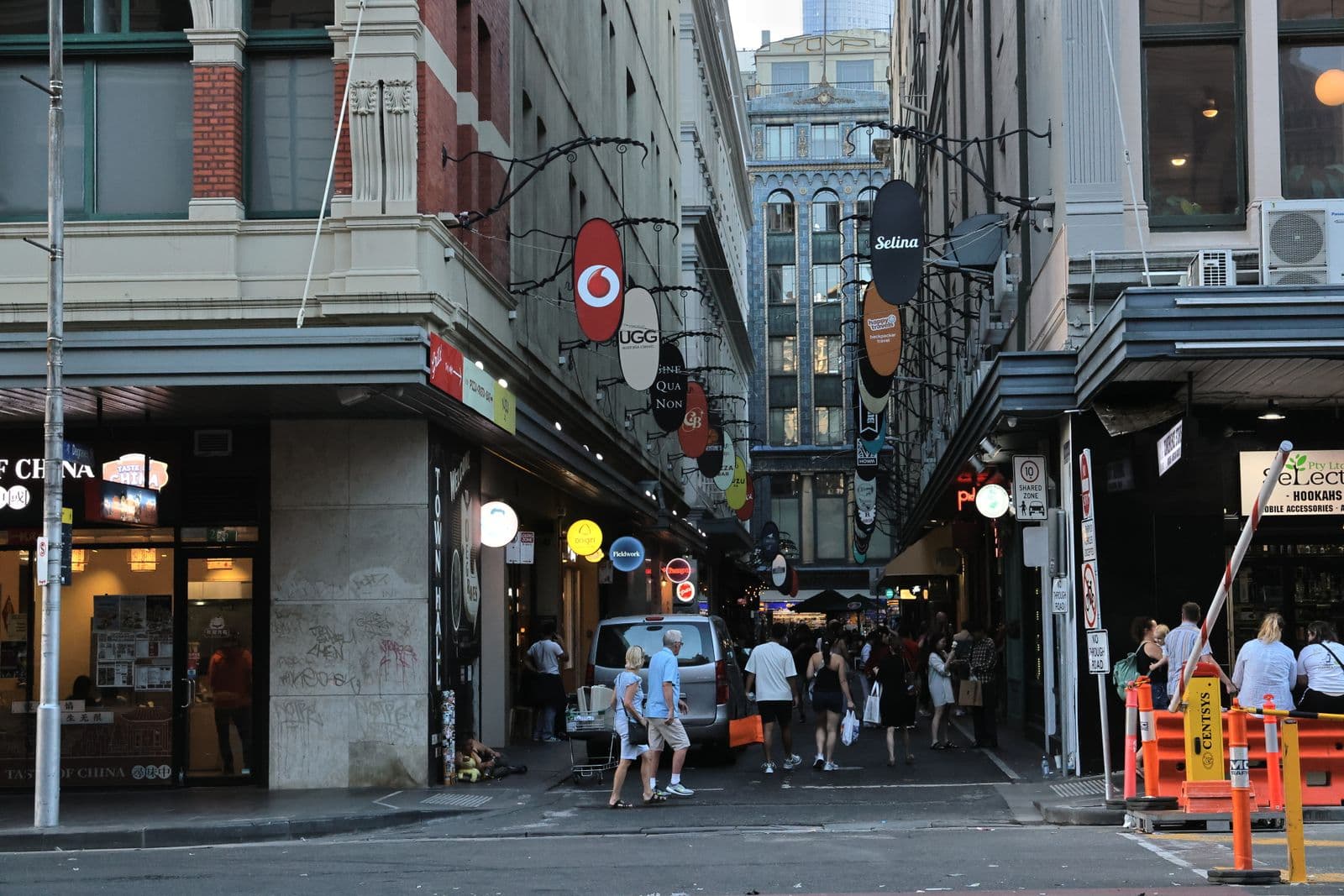 The mouth of a narrow city laneway, oval blade signs on wrought-iron brackets overhead, pedestrians walking in