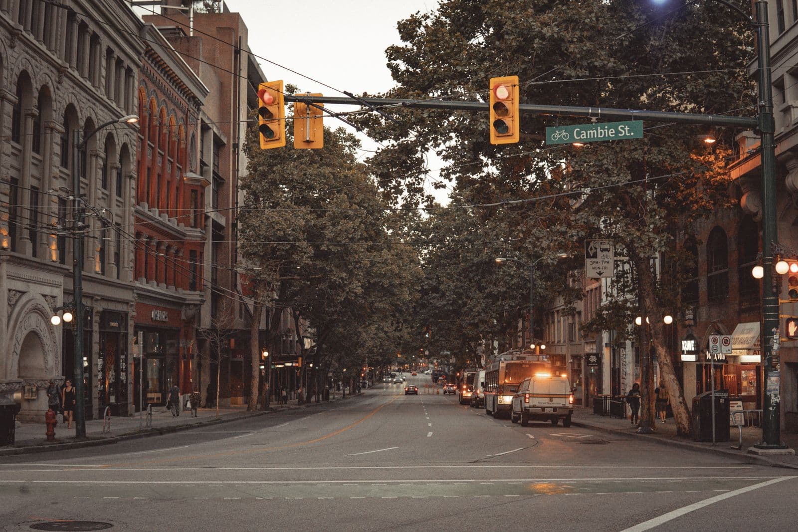 A quiet tree-lined street at dusk, heritage stone and brick frontages, yellow traffic lights, a parked utility truck