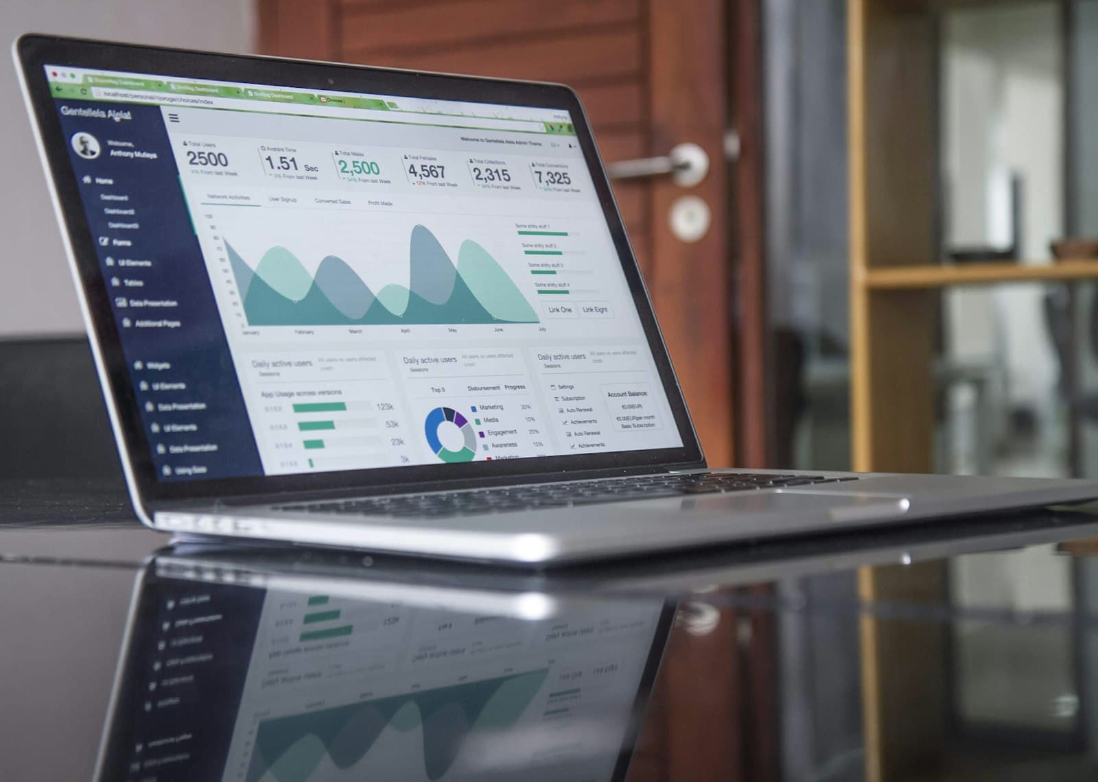 An open laptop on a dark glossy table showing an admin dashboard of user totals and traffic charts