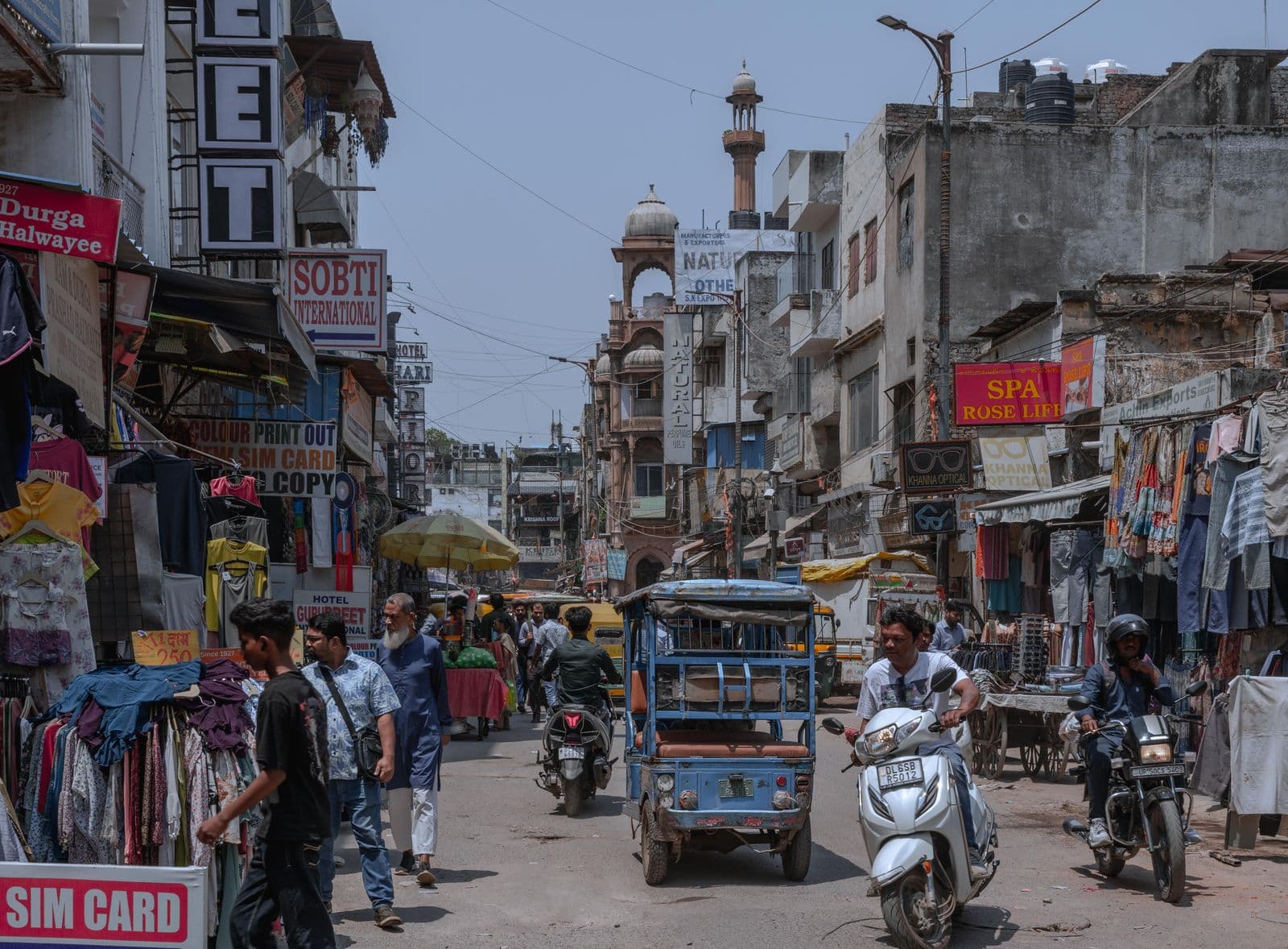 A busy market street in daylight, stacked painted shop boards, clothes on racks, an e-rickshaw and scooters