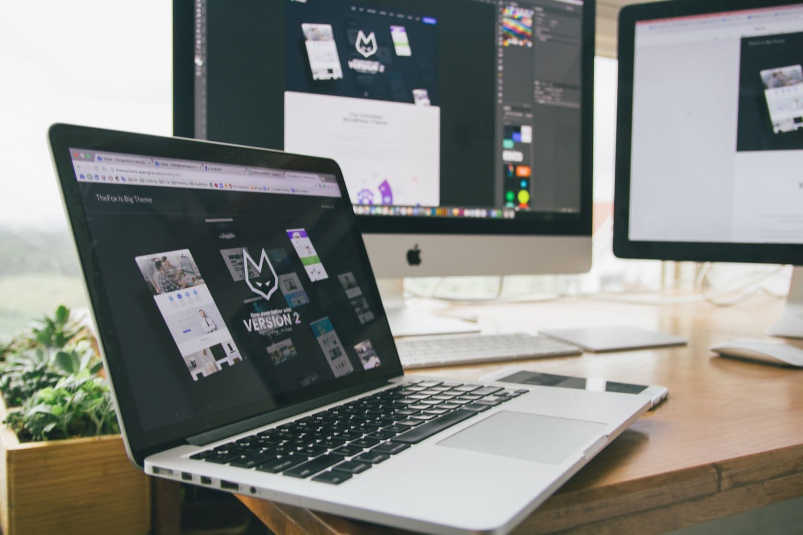 A laptop on a desk showing a website landing page, two larger screens behind it with the design open in an editor
