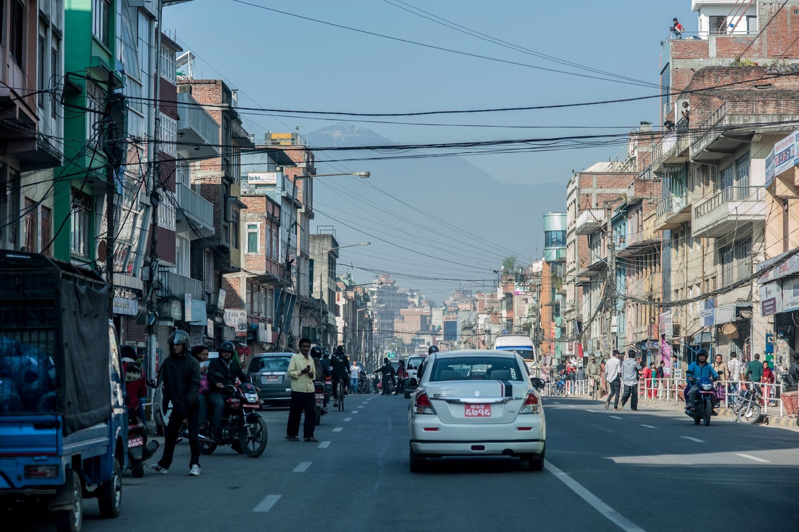 A busy Kathmandu commercial street with shophouses, tangled overhead power lines, motorbikes and a taxi