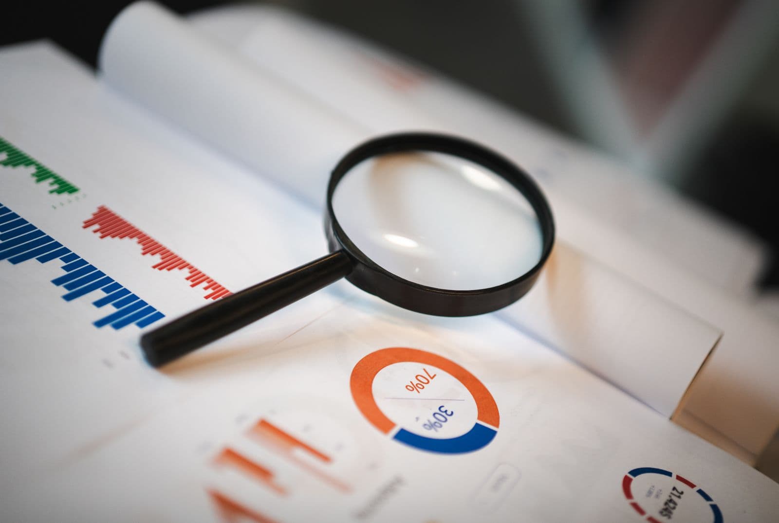A magnifying glass resting on printed reports showing bar charts and split donut charts