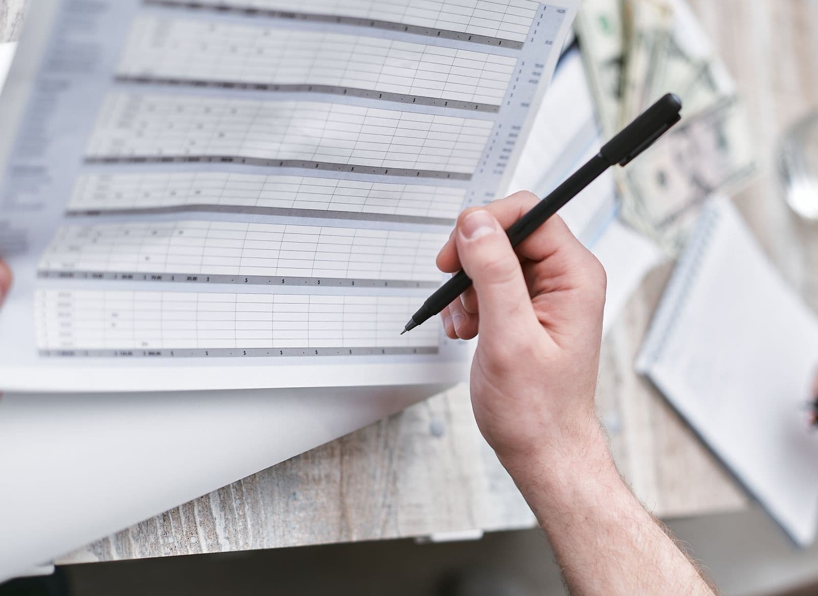 A hand holding a pen over printed spreadsheet pages of cost columns, banknotes on the desk beside them