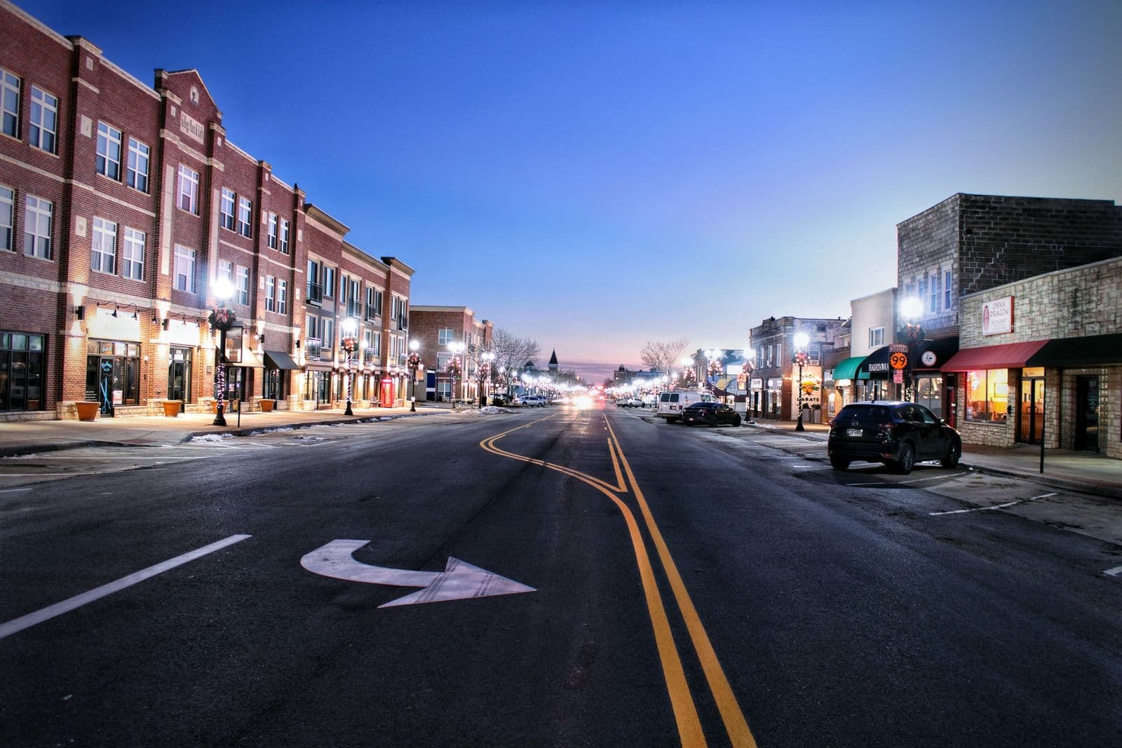A small-town main street at twilight, red-brick storefronts, lit lamp posts with wreaths, cars parked at the kerb