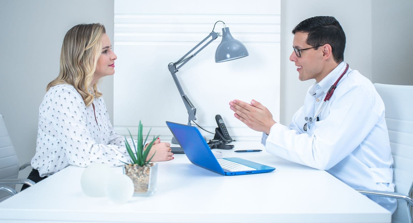 A doctor in a white coat talking with a seated visitor across a white desk with a laptop