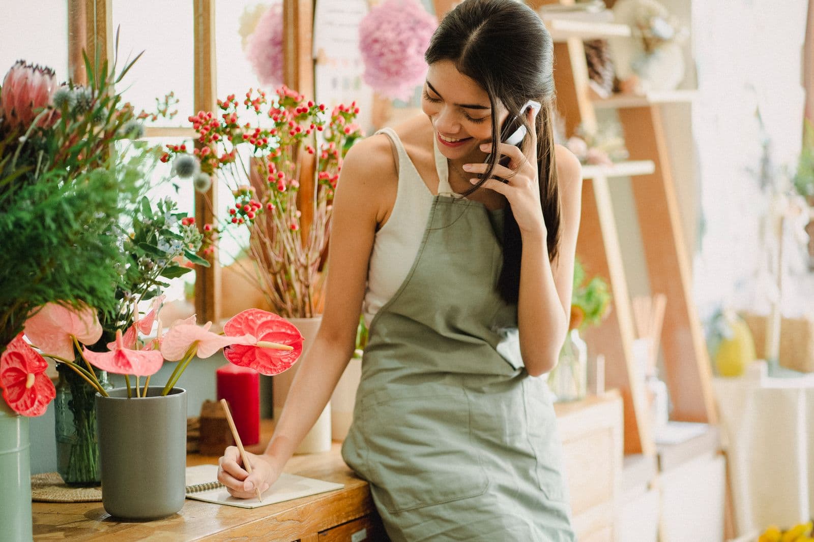 A florist in an apron taking a phone call in a flower shop, writing in a notebook on the counter