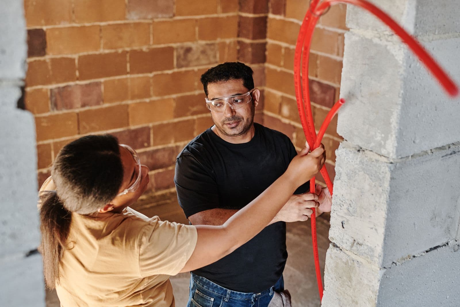 Two workers in safety glasses fitting red plastic pipe to a block wall inside an unfinished building