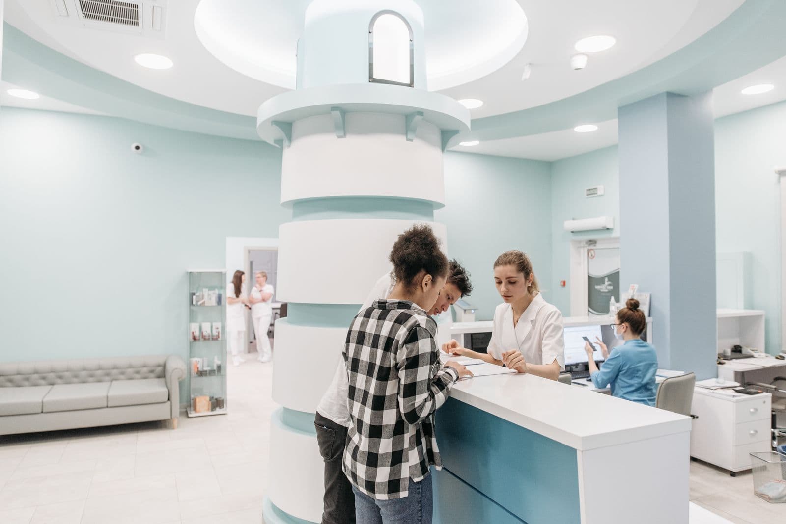 A clinic reception counter where a staff member in white attends to two people standing at the desk