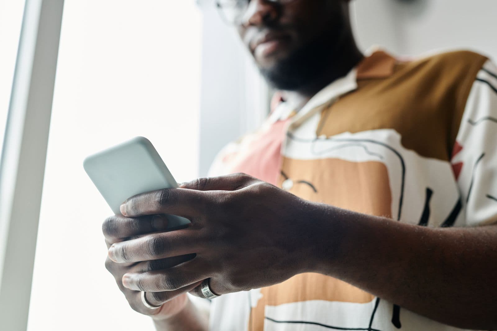 A man in a patterned shirt holding a smartphone in both hands beside a bright window