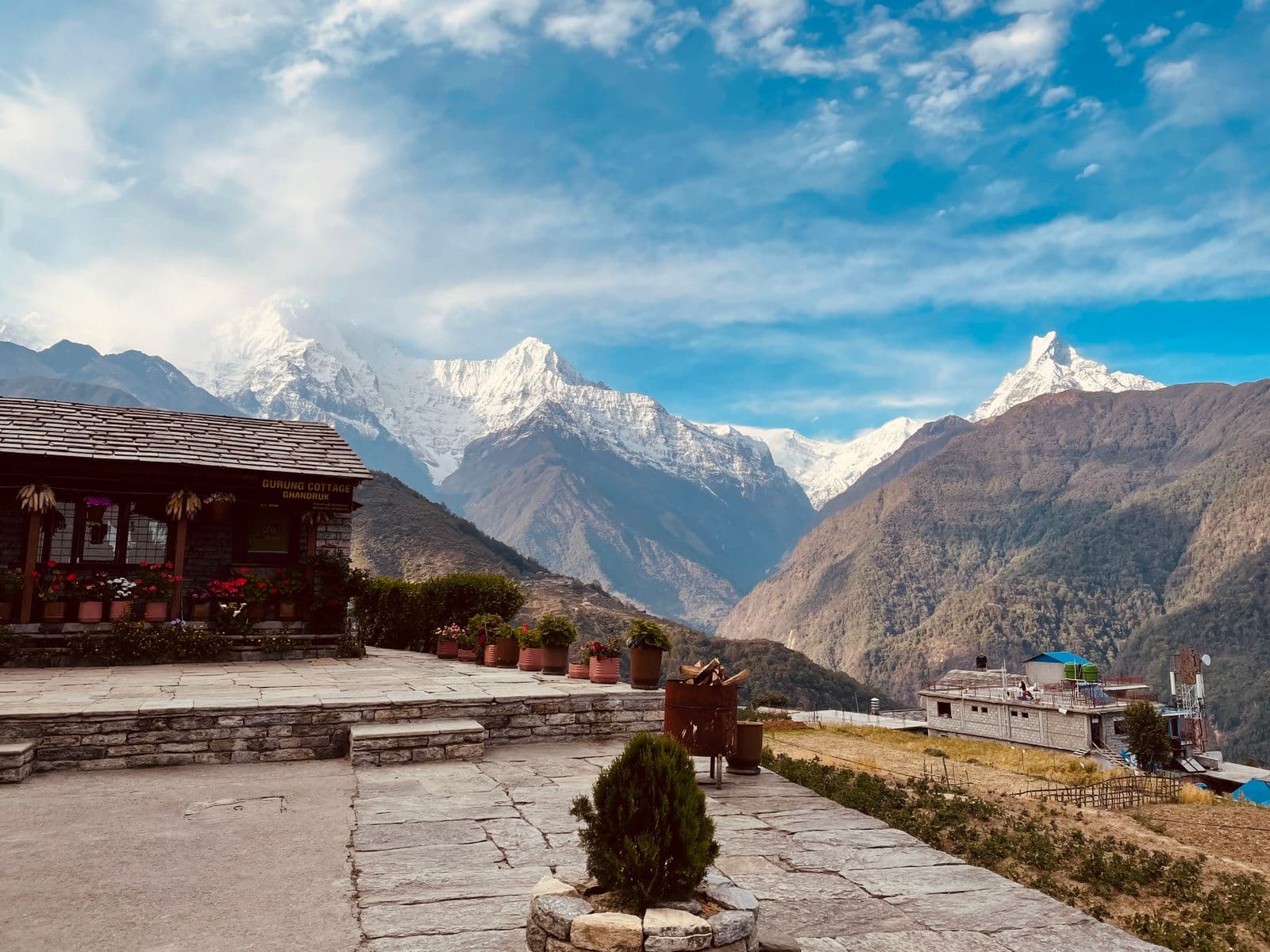 A stone-paved guesthouse terrace with potted flowers, snow-capped Himalayan peaks and a deep valley beyond
