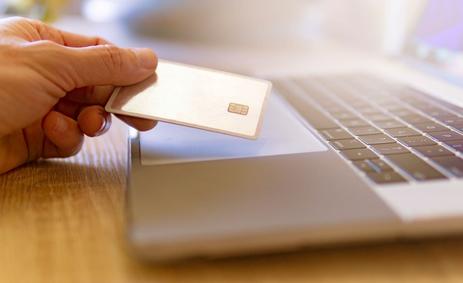 A hand holding a blank chip payment card above the trackpad of an open laptop on a wooden desk