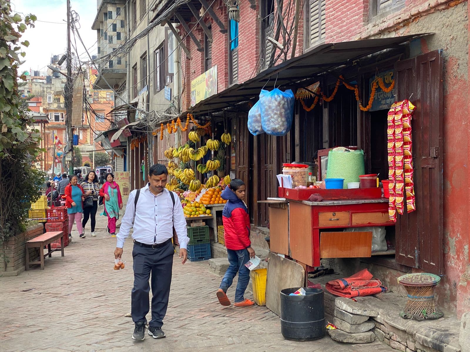 A brick-paved Kathmandu lane with an open-fronted fruit shop and a red street-food cart hung with marigold garlands