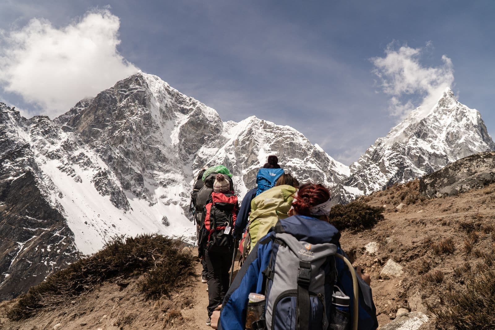 A line of trekkers with large backpacks climbing a dusty trail toward snow-covered Himalayan peaks