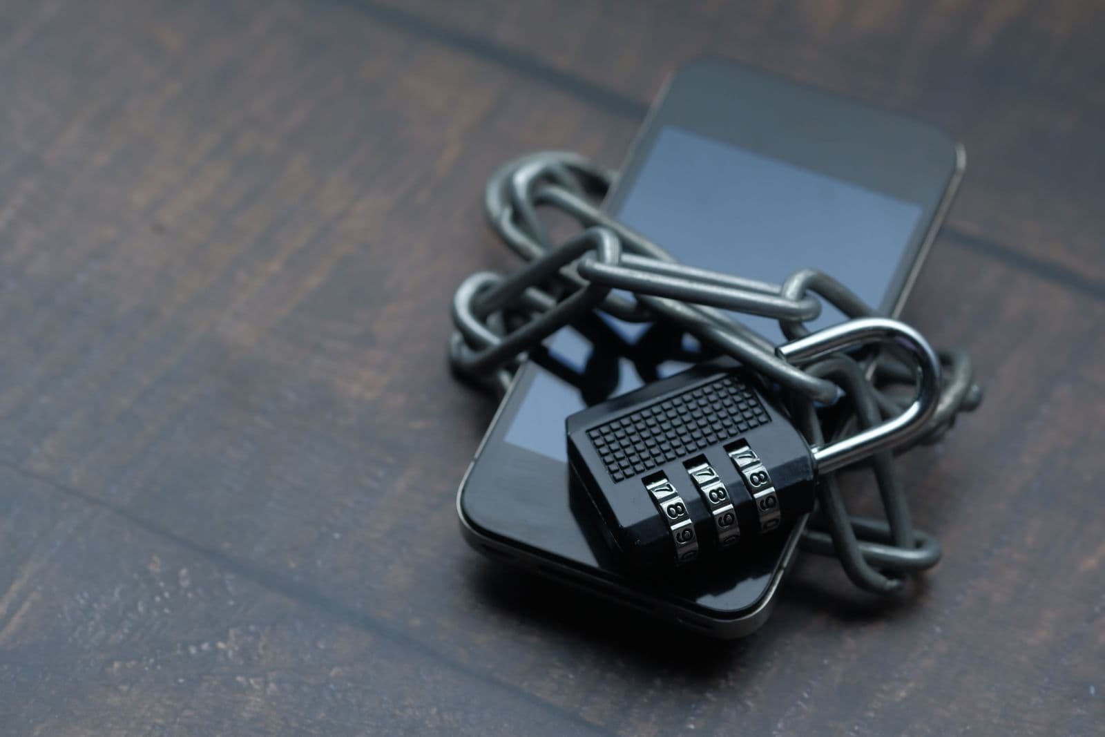 A smartphone on dark wood, wrapped in a heavy steel chain locked with a combination padlock