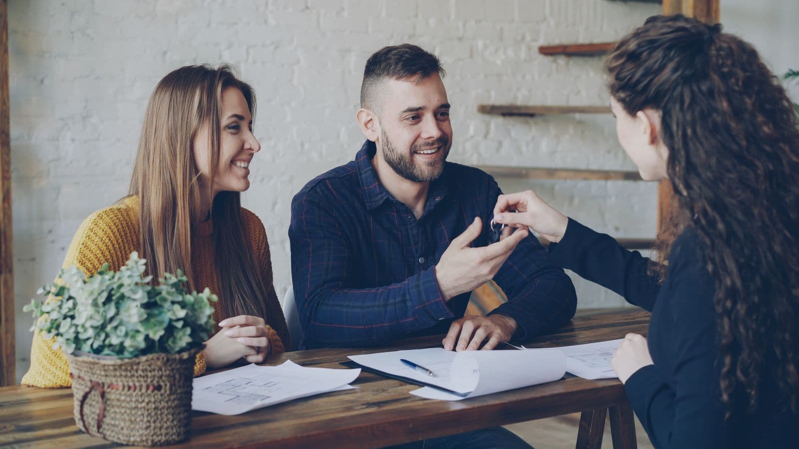 An agent handing keys across a table to a smiling couple, floor plans and a signed document between them