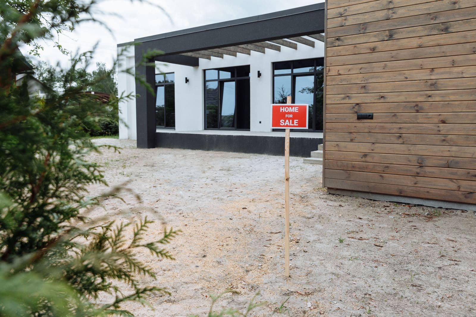 A red Home For Sale sign on a post in the sandy yard of a modern single-storey house