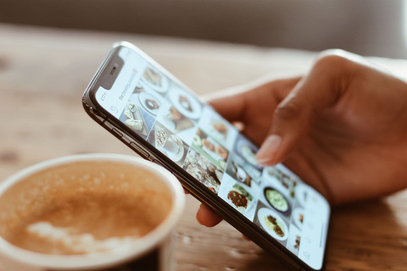 A hand holding a phone showing a grid of food photos, beside a coffee cup