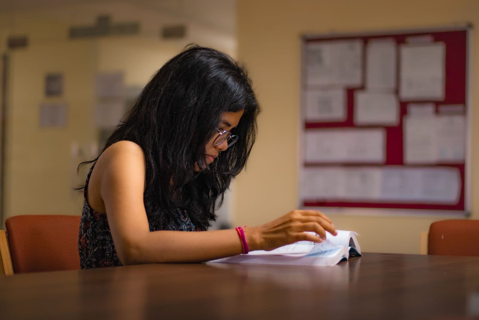 A student in glasses reading a thick book at a table, a paper-covered noticeboard on the wall behind
