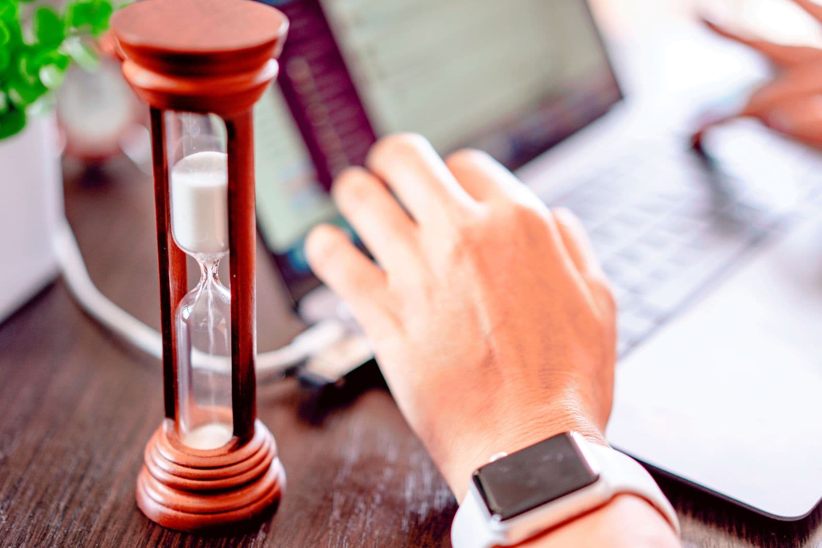 A wooden hourglass running on a desk, a hand reaching past it toward a laptop keyboard