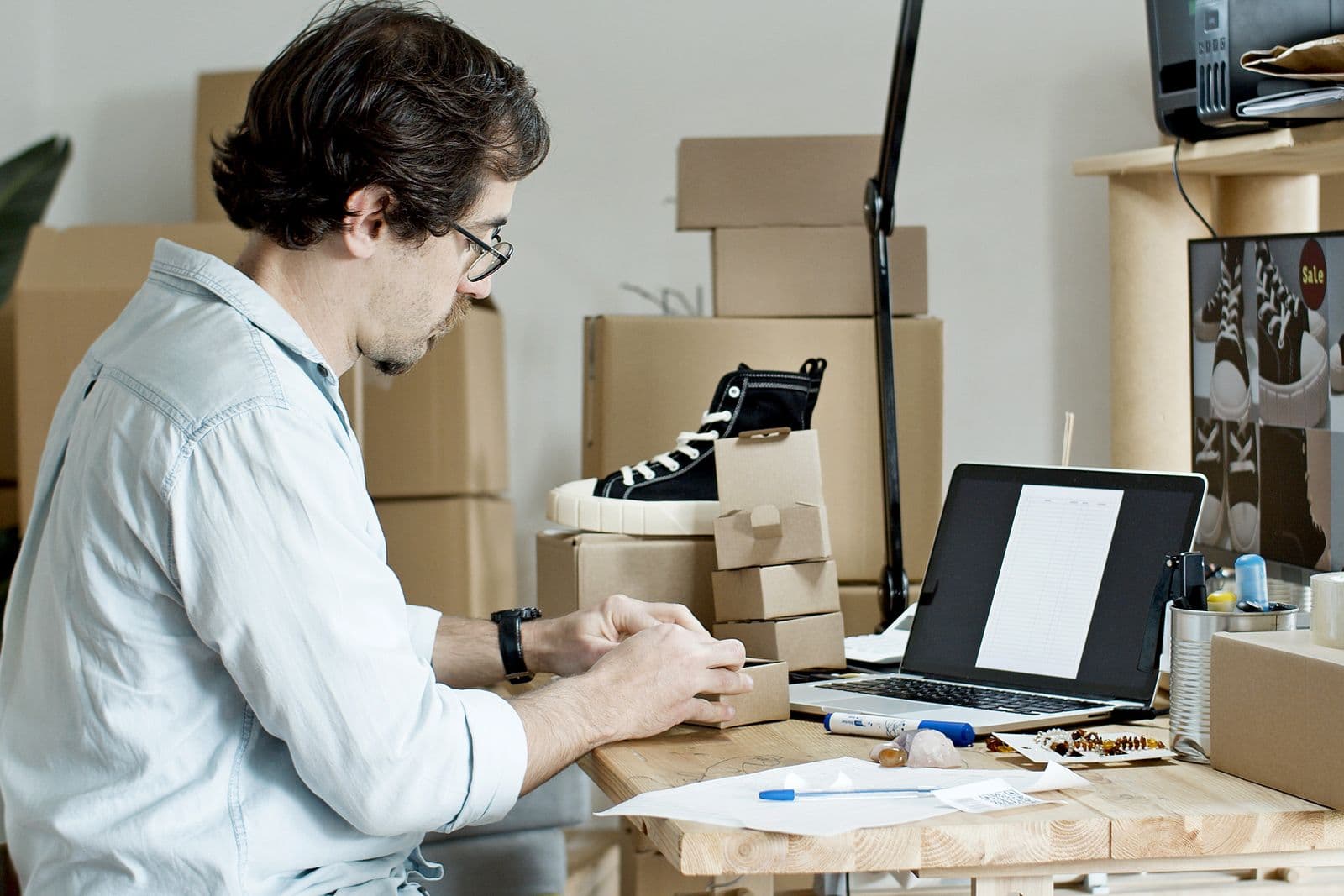 Man folding kraft shipping boxes at a bench beside a laptop, surrounded by stacked cartons