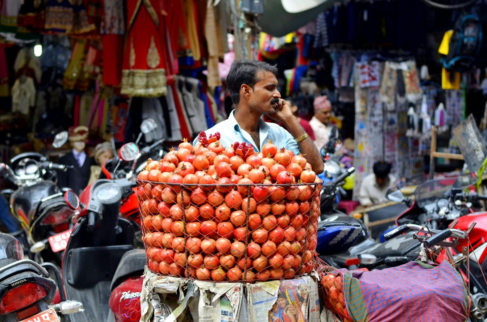 A fruit vendor on the phone behind a large basket of pomegranates at a market stall