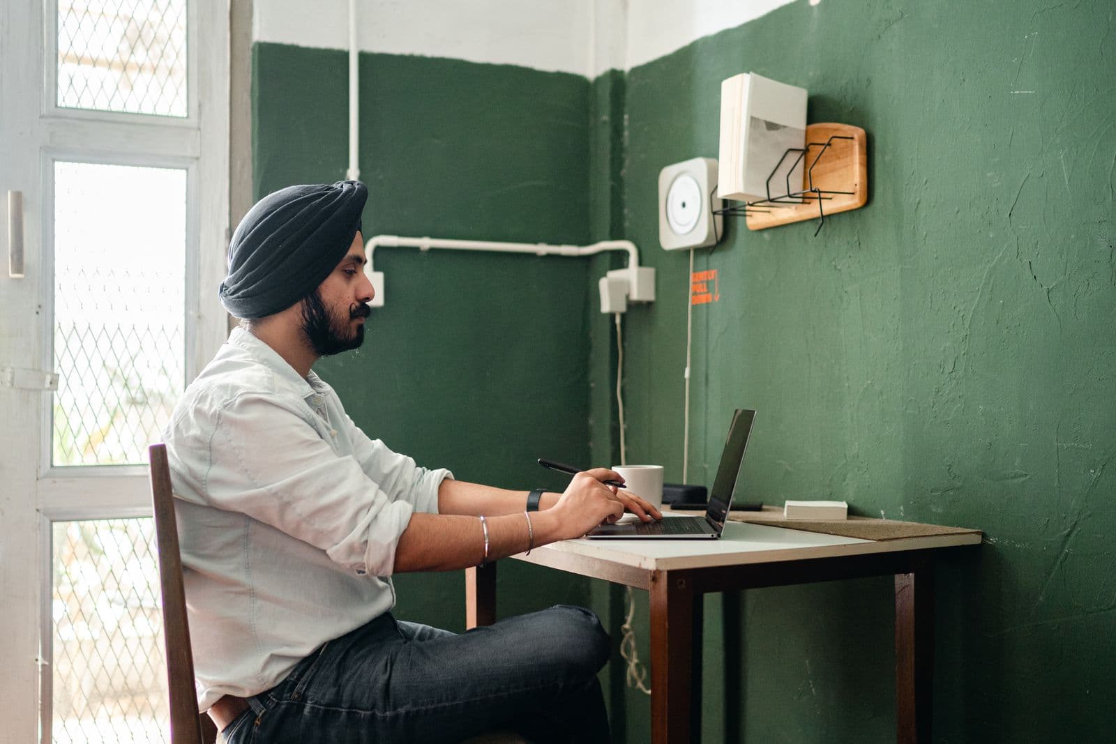 A man in a turban typing on a laptop at a small table in a plain room