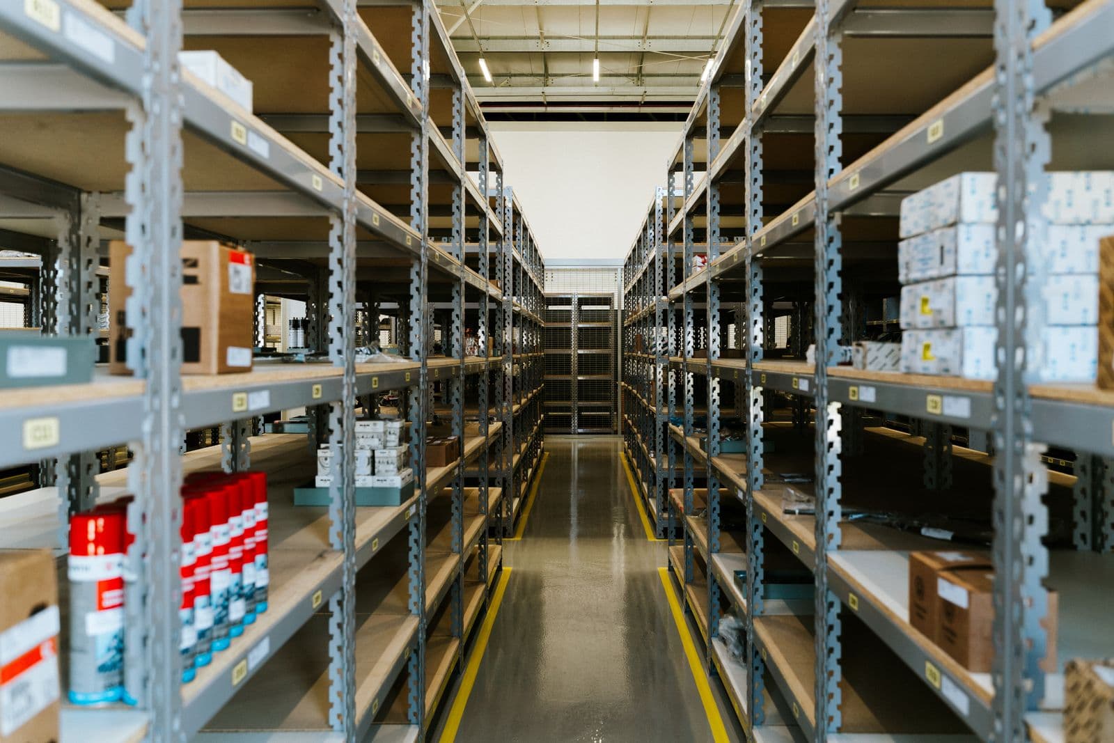 A stockroom aisle between grey steel shelves holding cardboard cartons and boxed goods